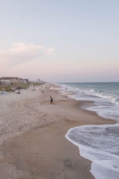 people are walking on the beach near the water and buildings in the distance, with waves coming up from the shore