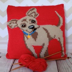 a red pillow with a dog on it next to some knitting needles and a ball of yarn