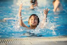 a young boy playing in a swimming pool with his hands up to the water as people swim behind him