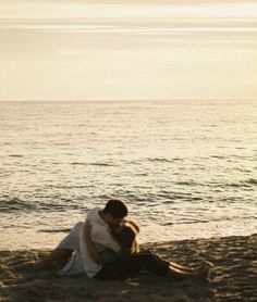 a man and woman sitting on top of a sandy beach next to the ocean at sunset