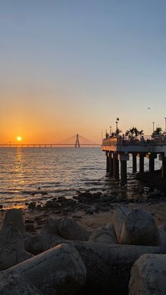 the sun is setting over the ocean with rocks on the shore and a pier in the distance