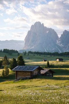 a small cabin in the middle of a grassy field with mountains in the background and clouds in the sky