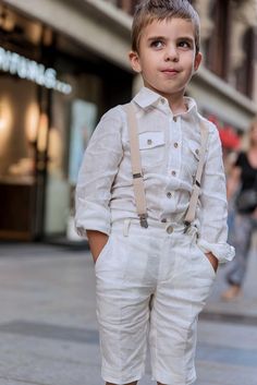 a young boy in white pants and suspenders standing on the sidewalk with his hands in his pockets