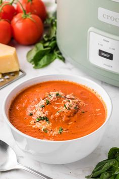 a white bowl filled with tomato soup next to a crock pot and some cheese