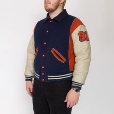 Vintage 70s letterman jacket from a high school team called the Terrors, in navy blue wool with off-white leather sleeves, orange color blocking, and striped knit trim. Lightly distressed. Measurements and Condition: Fits like: Labeled size 50, fits modern men's XL Fabric: Feels like a wool shell, leather sleeves, acrylic knit trim, and quilted nylon lining with polyester fill Brand: Ripon Jackets, made in USA Condition: Lightly distressed, with light general wear, some spots and discoloration on the sleeves, faint discoloration on the cuffs and middle of the patch on the back, and a few other minor scattered small faint spots. Length: 25.5" Chest: 52" Waist: 40" to 44" Shoulders (seam to seam): 23" Sleeve: 24" Shown on a 6'4" model, who usually wears a size extra large. See our FAQ for mo Varsity Letterman Jackets, Letterman Jacket, Leather Sleeve, Jacket Vintage, Blue Wool, Modern Man, Striped Knit, White Leather, Team Logo