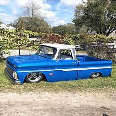 an old blue pickup truck parked in the grass next to a chain link fence and trees