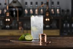 a tall glass filled with ice and lime next to a copper shaker on a wooden table