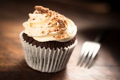 a cupcake with white frosting and chocolate shavings sits on a table next to a fork