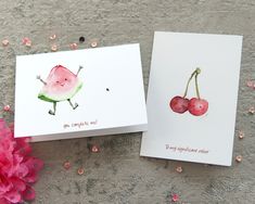 two cards with watermelon and cherries on them sitting next to pink flowers