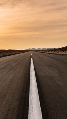 an empty road with white lines in the middle and mountains in the background at sunset