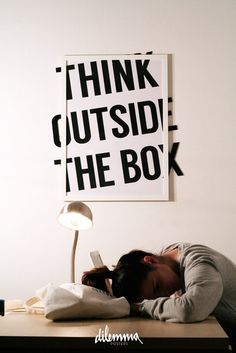 a person is sleeping on a desk with a lamp and poster behind them that says think outside the box