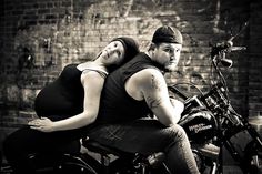 a man and woman sitting on a motorcycle in front of a brick wall, with their arms around each other