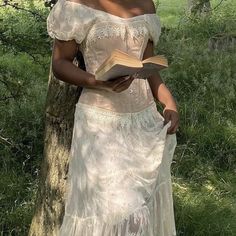 a woman in a white dress standing next to a tree holding a book and looking at the camera