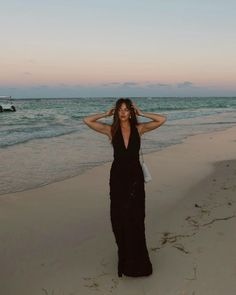 a woman in a black dress standing on the beach at sunset with her hands behind her head