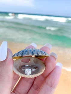 a person holding a shell with a diamond ring in it on the beach near the ocean