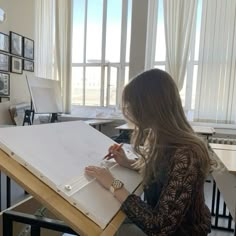 a woman sitting at a desk drawing on a piece of paper in front of a window