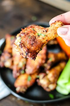 a hand holding up a piece of chicken wings over a black plate with carrots and celery