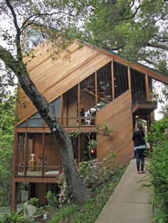 a woman walking up the side of a wooden house next to a tree and bushes