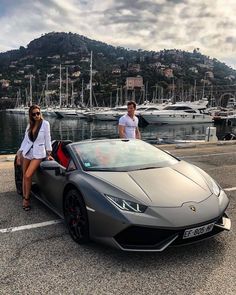 a man and woman standing next to a grey sports car in front of some boats