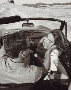 a man and woman sitting in the back of a convertible car on a dirt road