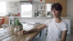 a woman sitting at a wooden table with a plant in a vase on top of it