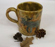 a coffee cup with an animal on it next to two pine cones and a leaf