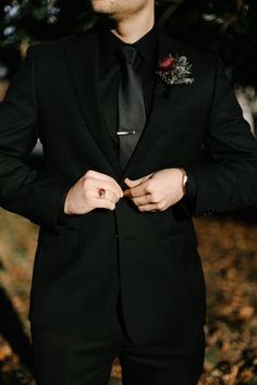 a man wearing a black suit and tie with his hands in his pockets while standing outside