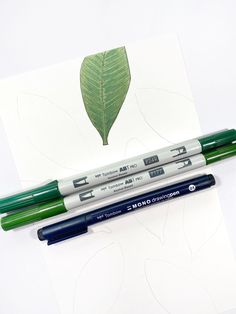 two pens sitting next to each other on top of a piece of paper with a green leaf