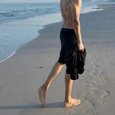 a shirtless man walking on the beach with his hand in his pocket while wearing swim trunks