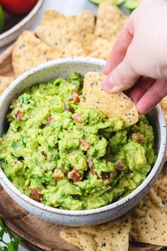 someone dipping guacamole into a bowl with tortilla chips and tomatoes in the background