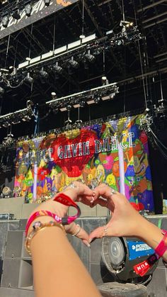 two people making a heart shape with their hands at an outdoor music festival in front of a stage