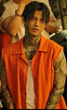 a man with tattoos on his neck and chest standing in front of other people wearing orange vests