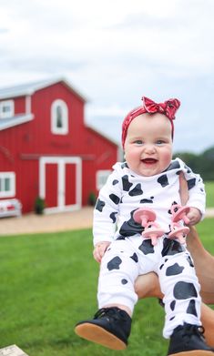 a woman holding a baby in front of a red barn with a cow print outfit