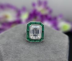 an emerald and diamond ring sitting on top of a gray surface with purple flowers in the background