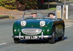 two people in a green convertible car driving down the street with another person behind them