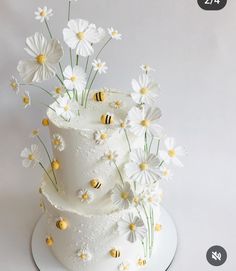 a three tiered white cake with daisies and bees on top, sitting on a plate