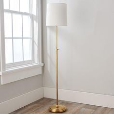 a floor lamp with a white shade on it in front of a window and wooden floor