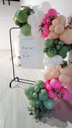 balloons and greenery are arranged around a sign for paint sip at an art gallery