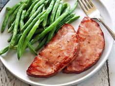 two slices of pork and green beans on a white plate with silverware next to it