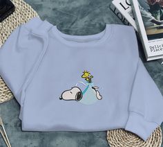 Embroidered Snoopy & Woodstock Sweatshirt  Wrap yourself in comfort and nostalgia with this beautifully embroidered sweatshirt featuring the beloved Snoopy and Woodstock from the Peanuts comic strip! This sweatshirt is perfect for casual days, chilly evenings, or simply relaxing at home. The detailed embroidery showcases Snoopy lounging in his signature laid-back style, while Woodstock adds a playful touch by balancing on his back with a kite-like string.  Available in various sizes, this sweatshirt is the perfect combination of nostalgia and comfort. Whether you're a lifelong Peanuts fan or just looking for a comfy sweatshirt with a cute design, this piece is sure to become a favorite in your wardrobe! Add this cozy, charming sweatshirt to your collection today! Cheap Pop Culture Cartoon Print Sweatshirt, Funny Snoopy, Snoopy Sweatshirt, Snoopy Sweater, Peanuts Comic Strip, Detailed Embroidery, Snoopy Woodstock, Dog Sweatshirt, Sweatshirt Cute