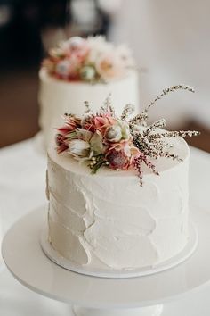 two white wedding cakes with flowers on each cake stand, one is frosted and the other is decorated