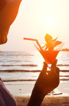 a person holding up a martini glass on the beach at sunset with an ocean in the background