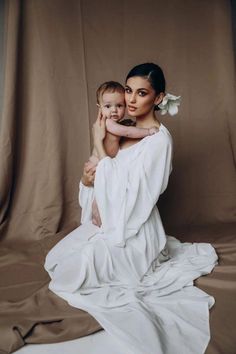 a woman holding a baby while sitting on top of a white cloth covered floor in front of a brown backdrop