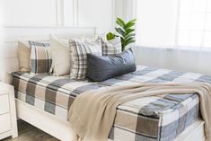 a bed with plaid sheets and pillows in a white room next to a window filled with plants