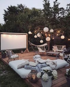 an outdoor movie screen and some chairs on a wooden deck with lights hanging from the trees