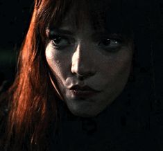 a woman with red hair and blue eyes stares at the camera while standing in front of a dark background