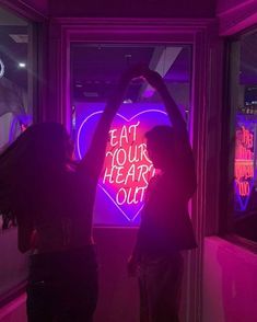 two people standing in front of a neon sign with the words eat your heart out