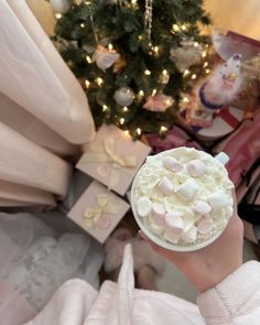 someone holding up a cup of marshmallows in front of a christmas tree