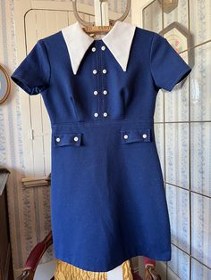 This cute dress comes from Wendy Lane, Montreal. It's made from fortrel in dark royal blue with a pointy wide fortrel collar and little white buttons down the front and on the faux pockets. It fastens with a long zipper in the back. The measurements, taken with the dress lying flat, are: shoulder to shoulder, 14 inches; armpit to armpit, 16 inches; sleeves, 7 inches; waist, 13 inches; length, 30 inches; bottom edge, 21 inches. In good condition. Fitted Blue Dress With Doll Collar, Blue Short Sleeve Dresses With Buttons, Fitted Collared Short Sleeve Dress, Preppy Fitted Knee-length Dresses, Fitted Preppy Mini Dress, Elegant Blue Dress With Doll Collar, Preppy Fitted Mini Dress, Blue Mini Dress With Buttons And Short Sleeves, Fitted Retro Dress With Doll Collar