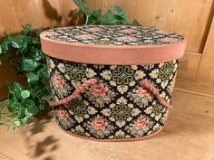 a small black and pink flowered box sitting on top of a wooden floor next to a plant